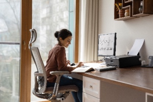 A young woman works on a computer in a modern home office, sitting in an ergonomic chair. The concept represents setting up a home office.